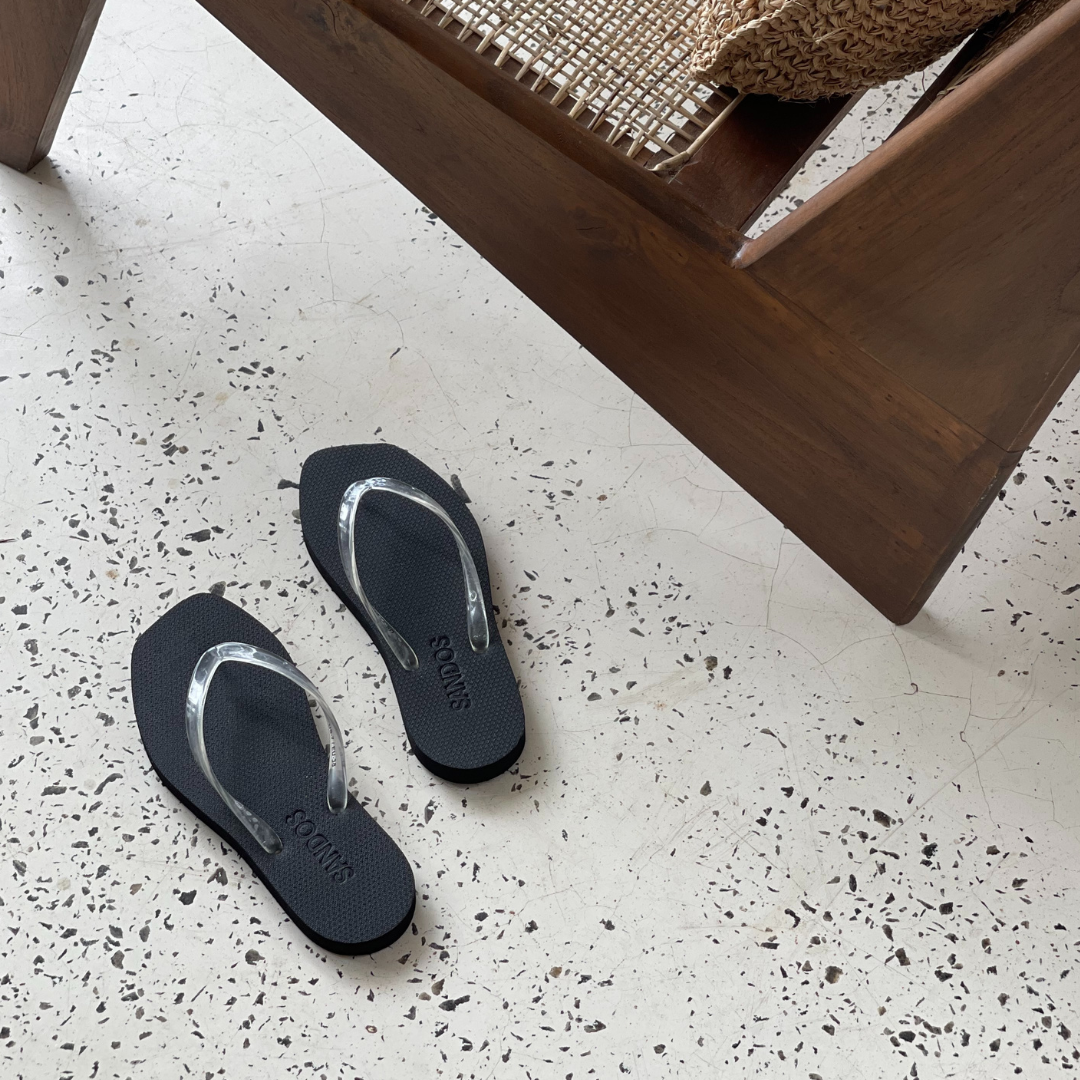 Sandos flip-flops with clear jelly straps on a speckled floor next to a wooden chair.