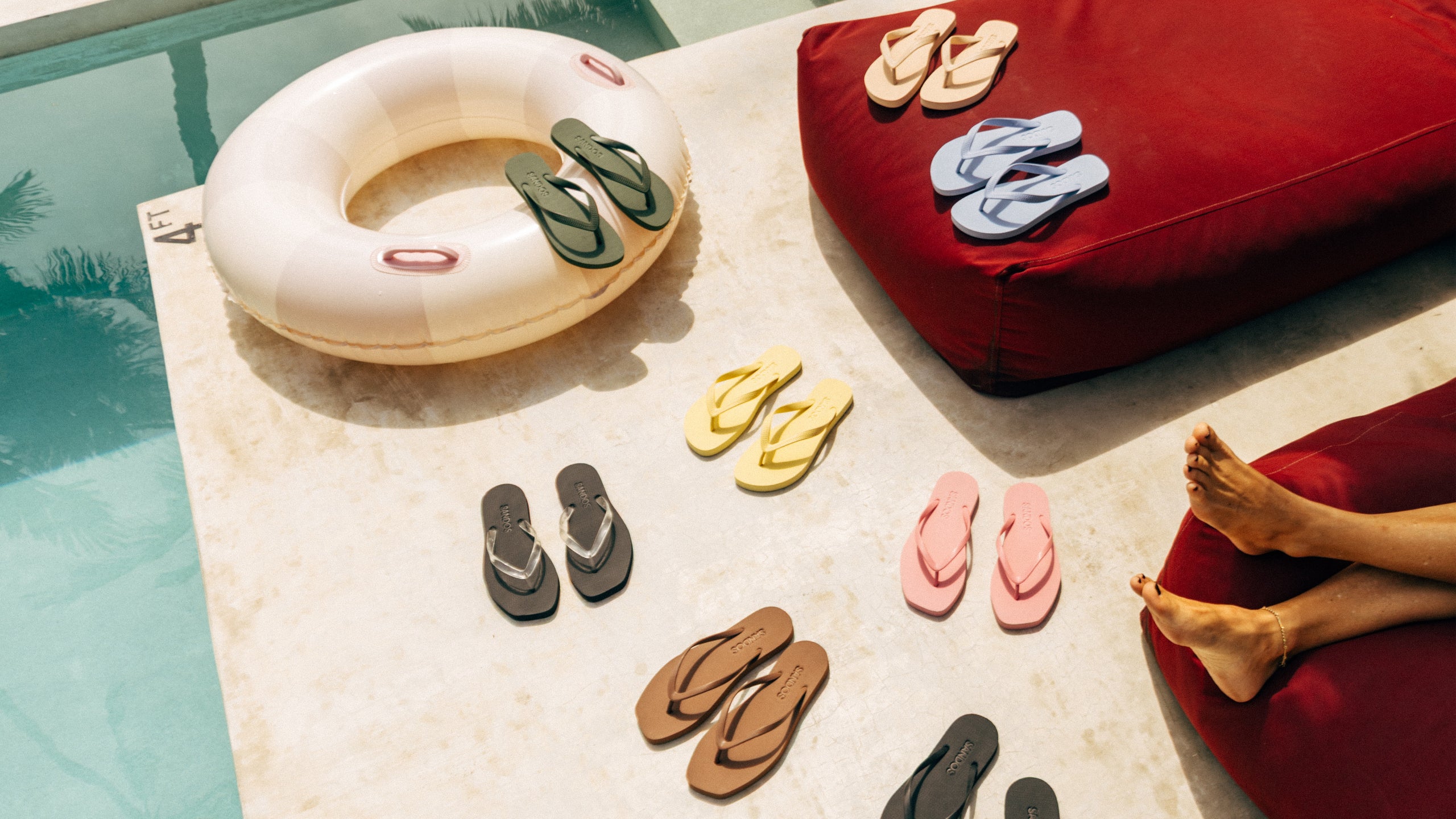 The range of Sandos flip-flops arranged on a poolside surface with a red cushion and inflatable ring.